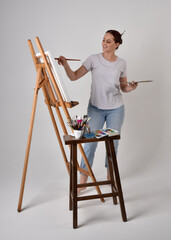 Wall Mural - Full length portrait of a red haired artist girl wearing casual jeans and white shirt.  standing pose 
painting a canvas on an easel, against a studio background.