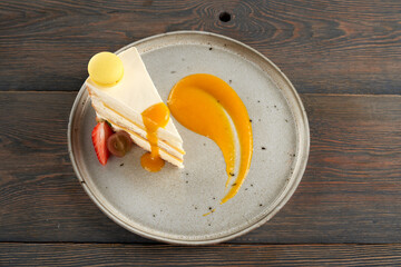 Top view of tasty orange and white cake on plate, isolated on wooden table. Delicious homemade slice of dessert served with berries, macaroon cookie and decorated with apricot puree.