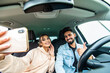 © F8  \ Suport Ukraine - road trip, leisure, couple, technology and people concept - happy indian man and woman driving in cabriolet car and taking picture with smartphone on selfie stick