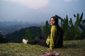 Kuala Lumpur, Malaysia - February 3rd, 2021 : Pretty girl sitting on grass, relaxing, enjoying Kuala Lumpur city view during sunset. Portrait of young Asian woman, wearing hijab and green attire.