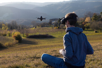 Young man wearing a VR glasses is piloting an fpv drone, he is sitting in a field at sunset. Image with selective focus.