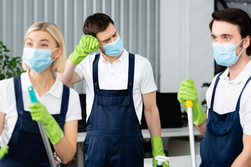 Wall Mural - Tired worker of cleaning service in medical mask standing near colleagues on blurred foreground in office