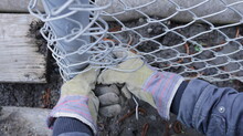 The Process Of Pulling A Galvanized Metal Mesh With Hands In Textile Gloves Around An Iron Post And Fixing The Mesh With Wire Top View, Installation Of A Metal Fence On An Outdoor Agricultural Plot