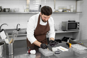 Wall Mural - Young male pastry chef in the process of making chocolates in a professional kitchen.The man holds a brush and carefully paints the candy molds with food coloring