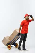 Delivery Concept - Portrait Of Handsome African American Delivery Man Or Courier Pushing Hand Truck With Stack Of Boxes. Isolated On Grey Studio Background. Copy Space.