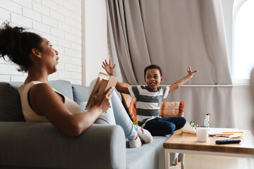 Black joyful woman reading book and having fun with her son at home