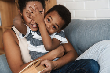 Wall Mural - Black smiling mother and son making fun and reading book on sofa at home