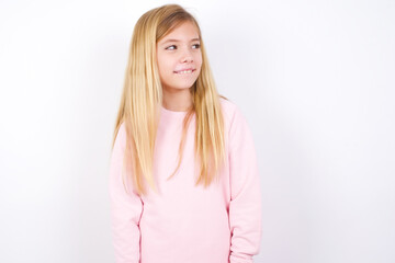 Wall Mural - beautiful caucasian little girl wearing pink hoodie over white background with thoughtful expression, looks away keeps hands down bitting his lip thinks about something pleasant.