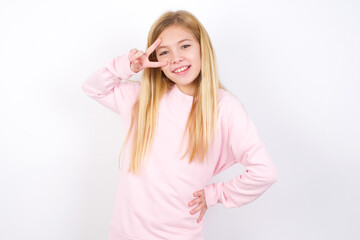 Wall Mural - beautiful caucasian little girl wearing pink hoodie over white background making v-sign near eyes. Leisure, coquettish, celebration, and flirt concept.