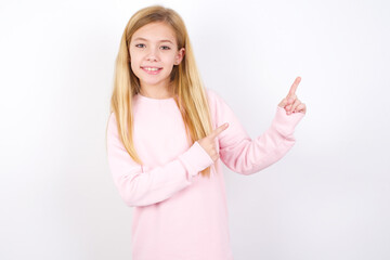 Wall Mural - Positive beautiful caucasian little girl wearing pink hoodie over white background with beaming smile pointing with two fingers and looking on empty copy space. Advertisement concept.