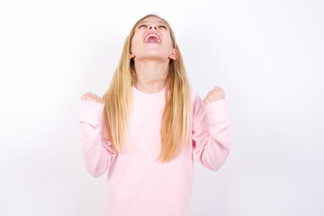 Wall Mural - beautiful caucasian little girl wearing pink hoodie over white background looks with excitement up, keeps hands raised, notices something unexpected.