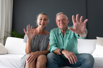 Happy caucasian senior couple in living room, on couch making video call, waving and smiling