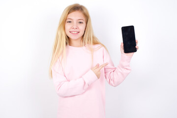 Wall Mural - Excited beautiful caucasian little girl wearing pink hoodie over white background holding and pointing with finger at smartphone with blank screen. Advertisement concept.