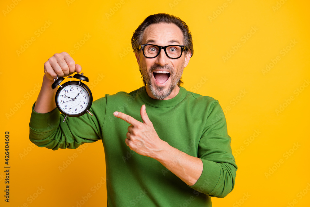 Photo of excited man hold alarm clock open mouth direct finger wear eyewear green sweater isolated yellow color background - obrazy, fototapety, plakaty 