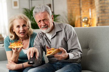 Wall Mural - Cheerful husband and wife sitting on sofa at home. Happy senior woman and man eating pizza while watching a movie.