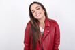 Positive young beautiful brunette woman wearing red biker jacket over white wall with overjoyed expression closes eyes and laughs shows white perfect teeth