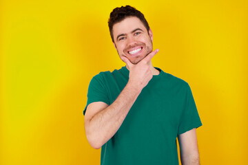 Wall Mural - young Caucasian man wearing green T-shirt against yellow wall looking confident at the camera smiling with crossed arms and hand raised on chin. Thinking positive.