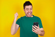 © Jihan - charming cheerful young handsome caucasian man wearing green t-shirt against yellow background holding in hands cell reading browsing