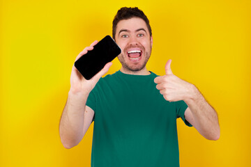 Wall Mural - Portrait young handsome caucasian man wearing green t-shirt against yellow background holding in hands cell showing giving black screen thumb up