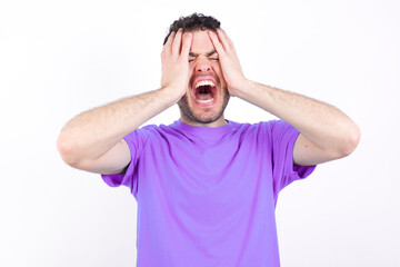 Shocked panic young handsome caucasian man wearing purple t-shirt against white background holding hands on head and screaming in despair and frustration.