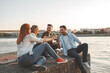 © BalanceFormCreative - Group of young people sitting by the river .They joying in city view . Young man playing guitar while his friends drinks beer .