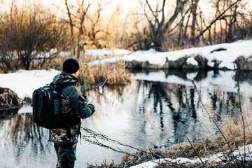 A fisherman with a fishing rod catches fish on the bank of a snow-covered river in early spring