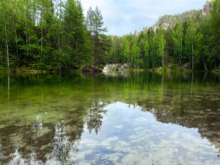 The green forest is reflected in the water. Rock on the lake shore. Summer landscape