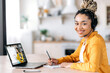© Kateryna - Distance learning by video call. Happy African American female student wearing headset, sits at work desk at home, studies remotely, looks at the camera, friendly smile,female teacher on laptop screen