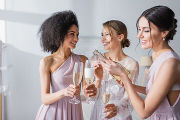 Poster - smiling woman pouring champagne near happy bride and african american bridesmaid.