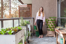 A Young Blonde Woman Is Planting A Vertical Tower Garden With Herbs And Vegetables On Her Urban Apartment Patio, In The Early Spring. She Is Holding A Watering Can, Ready For Gardening Season