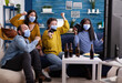 © DC Studio - Multiethnic friends celebrating victory while playing video games wearing face mask and keeping social distancing during global pandemic with corona in home living room raising hands.
