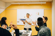 © Maskot - Smiling female entrepreneur showing numbers with thumbs up symbol to colleagues during meeting in office