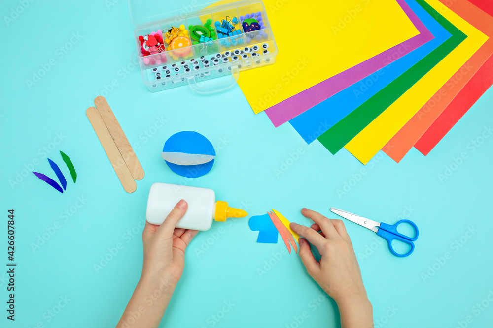 A child glues colored blue paper with glue at the table. Paper crafts for kids. - obrazy, fototapety, plakaty 