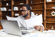 Clever and concentrated African-American guy is doing paperwork, a man looking through workpaper sitting at the desk with a laptop in office, a man checking report, collate data