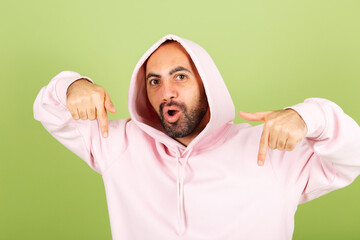 Young bald caucasian man in pink hoodie isolated on green background positive shocked amazed  excited point fingers down