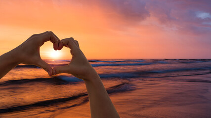 Wall Mural - Forming a heart shape with both hands in front of a beautiful sunset at the sea