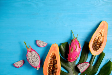 Wall Mural - Exotic fruit composition with monstera palm leaf. Papaya and pitaya, low-calorie nutritious fruits, whole and halved, visible pulp texture on blue wooden background. Close up, copy space, top view.