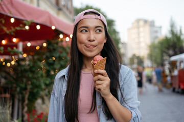 Portrait of beautiful asian woman eating ice cream on the street.  Emotional hipster wearing casual clothing holding tasty summer dessert looking away outdoors. Food festival