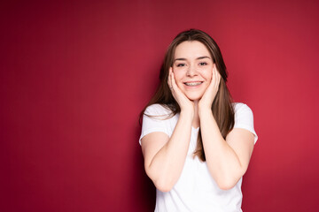 happy woman in braces and in a white t-shirt smiles broadly, touching her cheeks with her hands