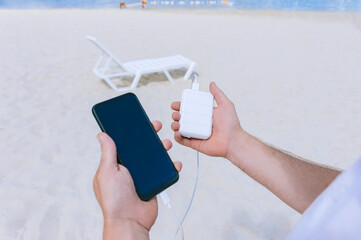 Wall Mural - Power Bank Energy charges the phone into the hands of a man on the beach. Against the backdrop of sand and a sun lounger.