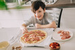 © Olga Krivokoneva - Little kid boy making pizza sitting at the table on the kitchen. Children helping in cooking lifestyle image