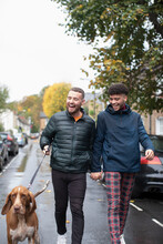 Happy Gay Male Couple Walking Dog On Wet Urban Street