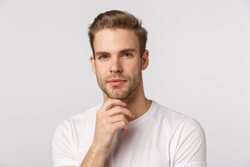 Hmm let me think. Pensive good-looking blond young bearded man in white t-shirt, touching chin and squinting intrigued, thinking, pondering what choice make, standing thoughtful