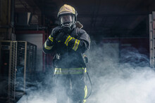 Portrait Of A Fireman Wearing Firefighter Turnouts And Helmet. Dark Background With Smoke And Blue Light.