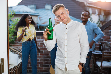 Happy young man in stylish outfit posing on camera with bottle of beer in hand. Three multiracial people dancing and talking behind. Rooftop party.