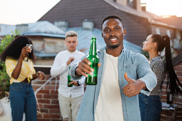 Wall Mural - Handsome african man in denim shirt holding beer and showing thumb up while three diverse friends dancing behind. Four friends hanging out together on open terrace.