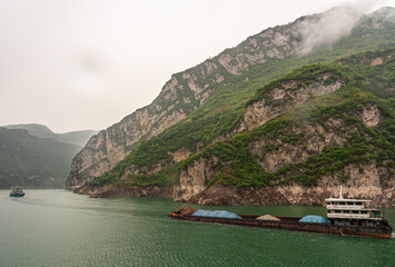 Yangtze River, Three Gorges, China - May 6, 2010: Zigui region. two barges sail on green water in front of green forested hills and mountains with beig-brown cliffs on shoreline under foggy sky.