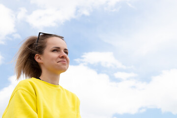 Smiling and happy young woman in yellow long sleeve stands under blue sky and white clouds. Portrait of adult girl. Positive mood and thinking.