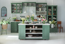 Green Kitchen Interior With Furniture. Stylish Cuisine With Flowers In Vase. Wooden Kitchen In Spring Decor. Cozy Home Decor. Kitchen Utensils, Dishes And Plate On Table. Kitchen Island In Dining Room