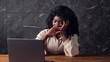 © lenblr - Exhausted black businesswoman with curly hair types on grey laptop lazily and yawns sitting at brown wooden table in office in late evening